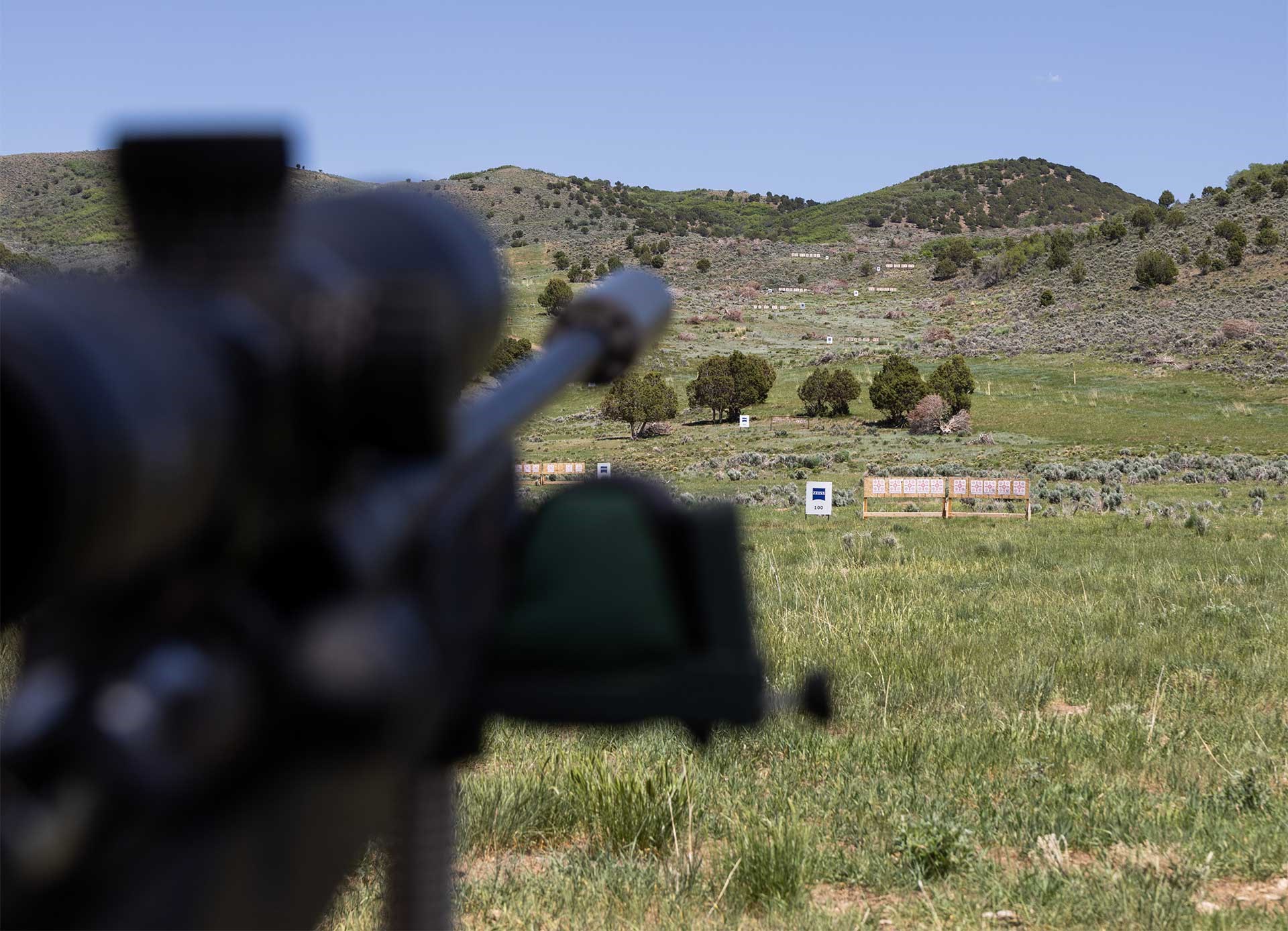 A rifle barrel and scope are seen in the foreground, while targets dot a hillside in the background.
