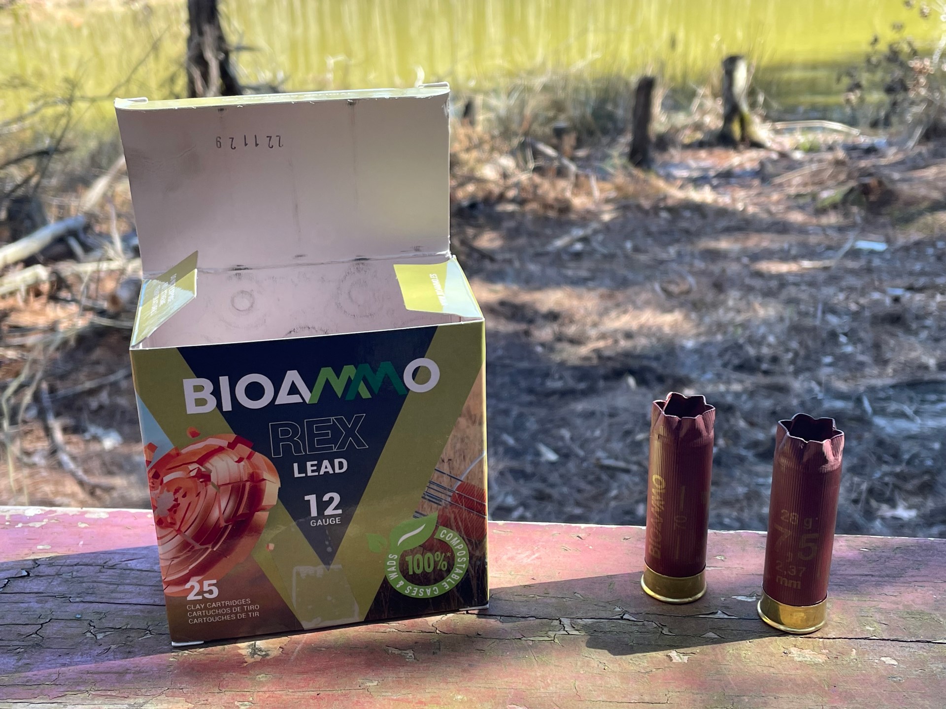BIOAMMO REX LEAD text on shotshell ammunition box two spent hulls outdoors stumps grass background