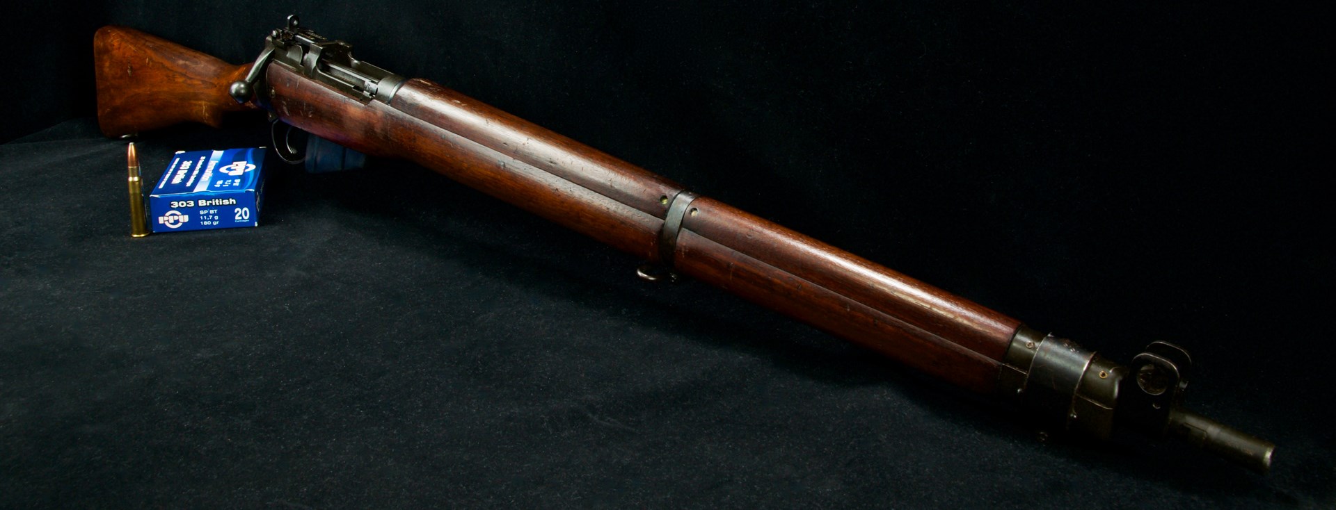 dynamic quarter view of lee-enfield bolt-action military surplus rifle black background shown with blue box of .303 British ammunition