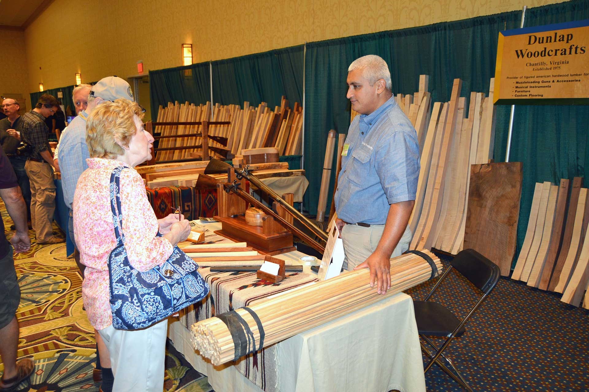 A pile of unfinished rifle stocks lay against the back wall behind a vendor and a woman talking at the CLA Show.