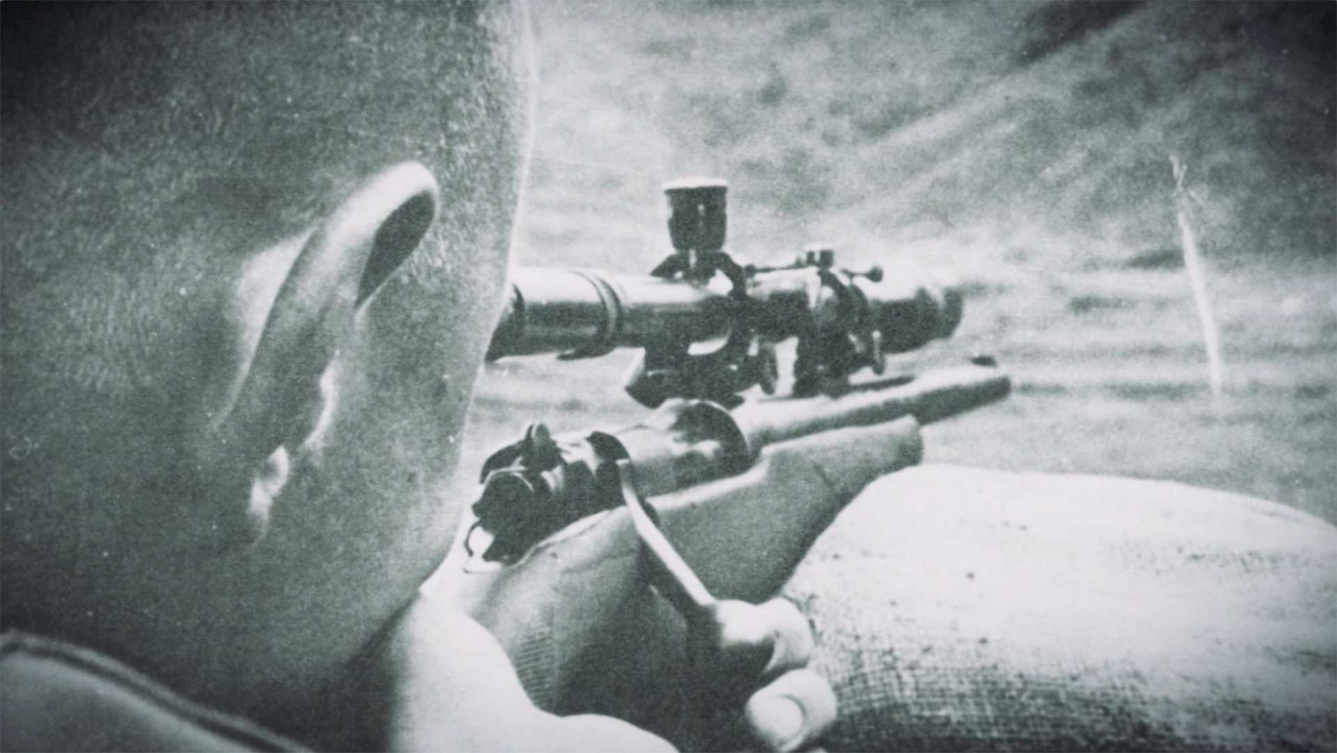 A Vietnam-era sniper in a black-and-white photo aims a scoped rifle into an open valley.
