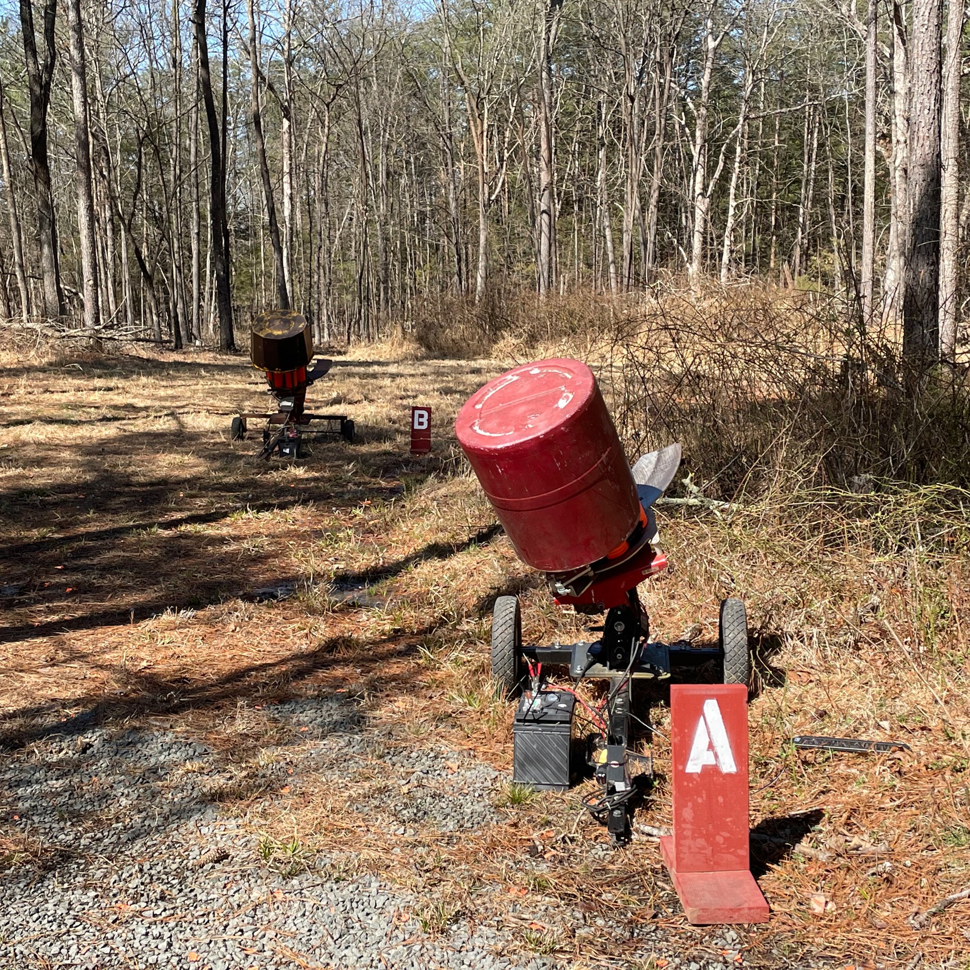 A and B clay target machines throwers shown from behind in forest setting shadows trees leaves grass gravel