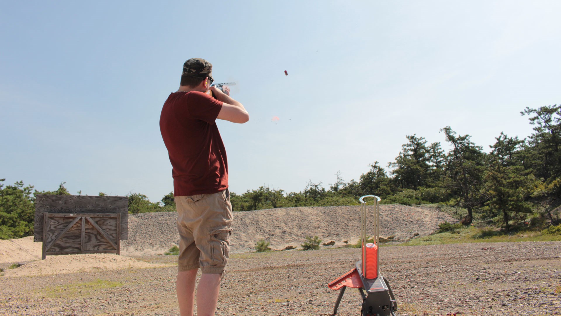 Man standing outdoors facing away from camera holding shooting shotgun with clay target machine blue sky dirt hill trees