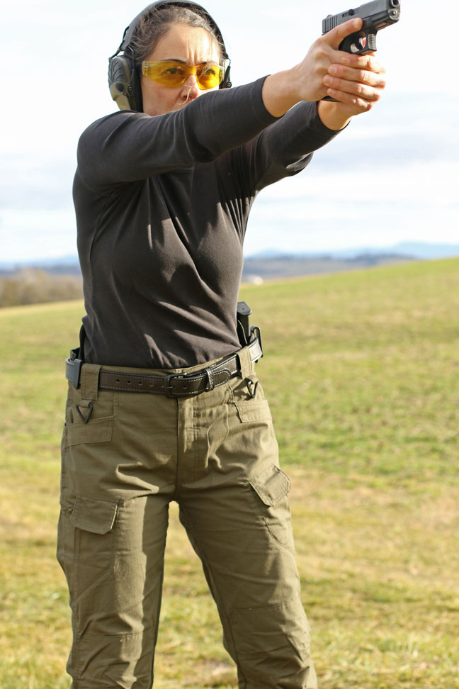 Forward-facing view of woman standing outstretched with pistol in hand and protective shooting gear on.