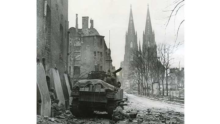G.I.s shelter behind an M4 Sherman tank on a shattered street in Cologne. The famous Gothic cathedral rises up in the distance.