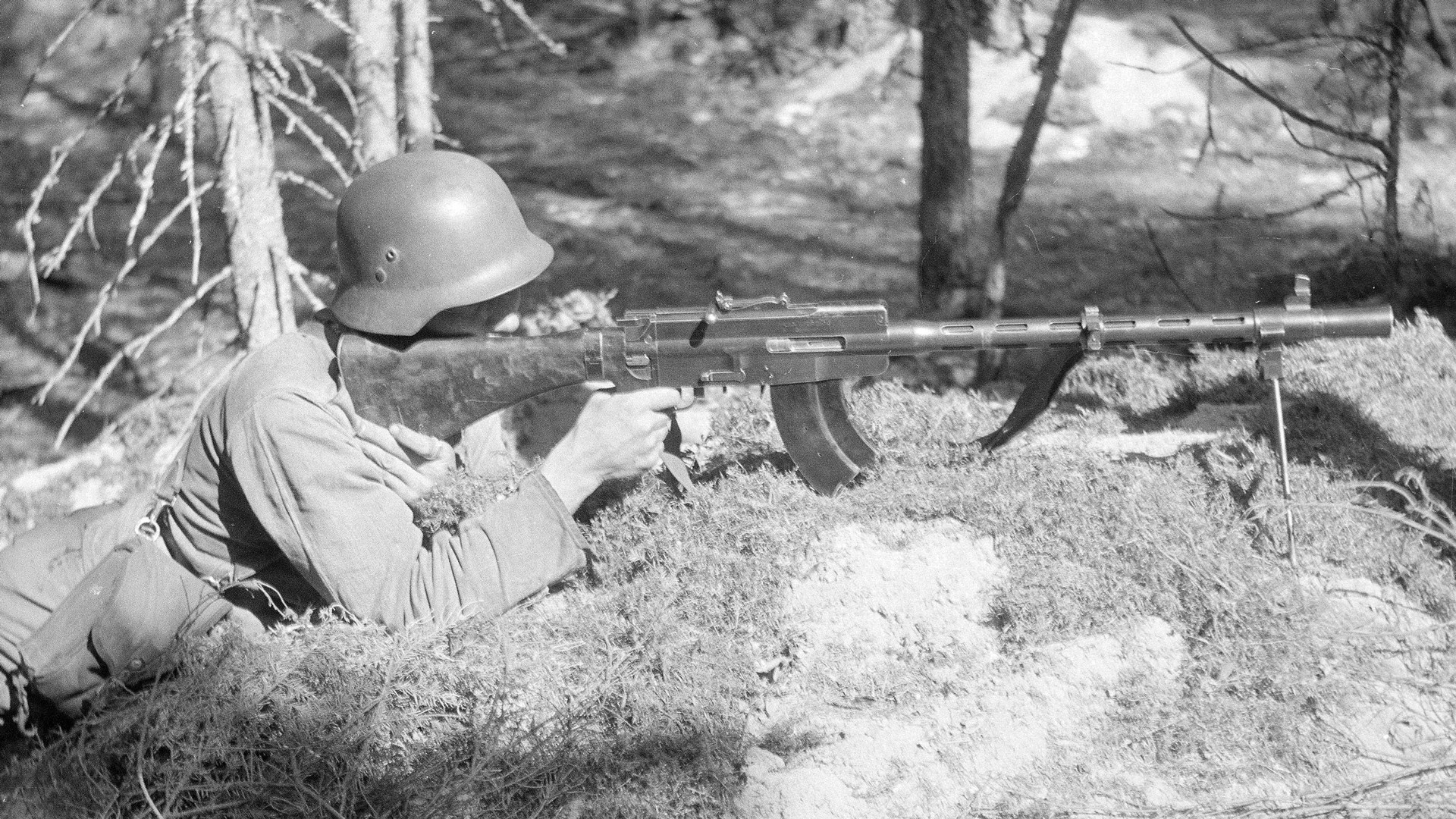 M/26 light machine gun right-side full-length profile view on bipod with finnish soldier aiming outdoors in woods on ground