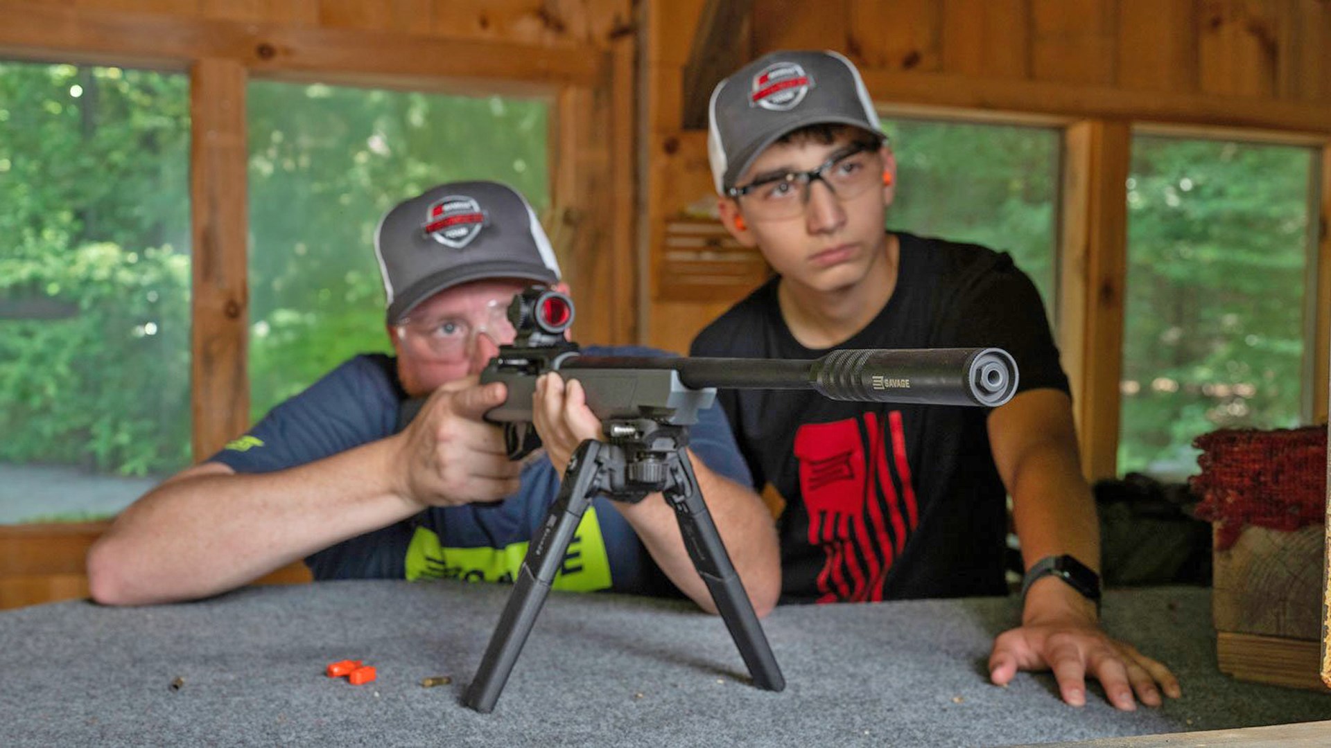 Two men aiming a Savage Arms RXR22 rifle off a shooting bench.