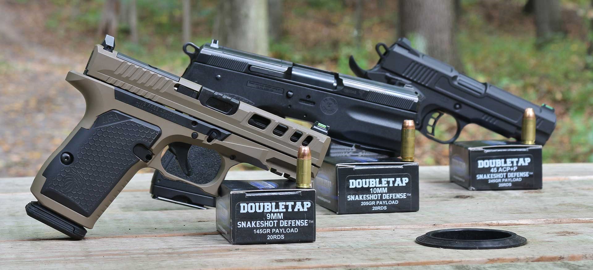 Three handguns propped up on a wood surface next to DoubleTap SnakeShot Defense ammunition boxes.