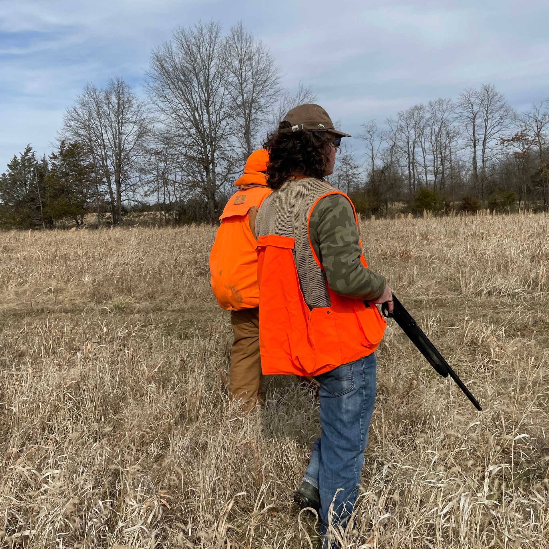 Two hunters in grassy field bird hunting shotgun muzzle direction safety rules blaze orange vest blue sky clouds trees black gun