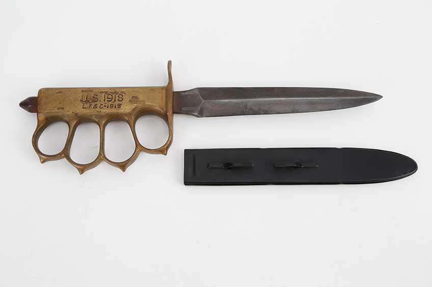 U.S. 1918 fighting knife with brass handle and brass finger guards on white background with sheath resting nearby.
