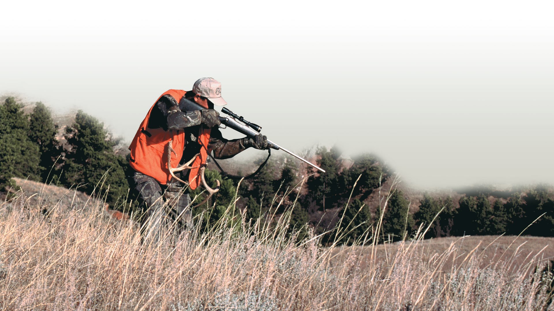 man in field with orange vest shooting rifle downhill