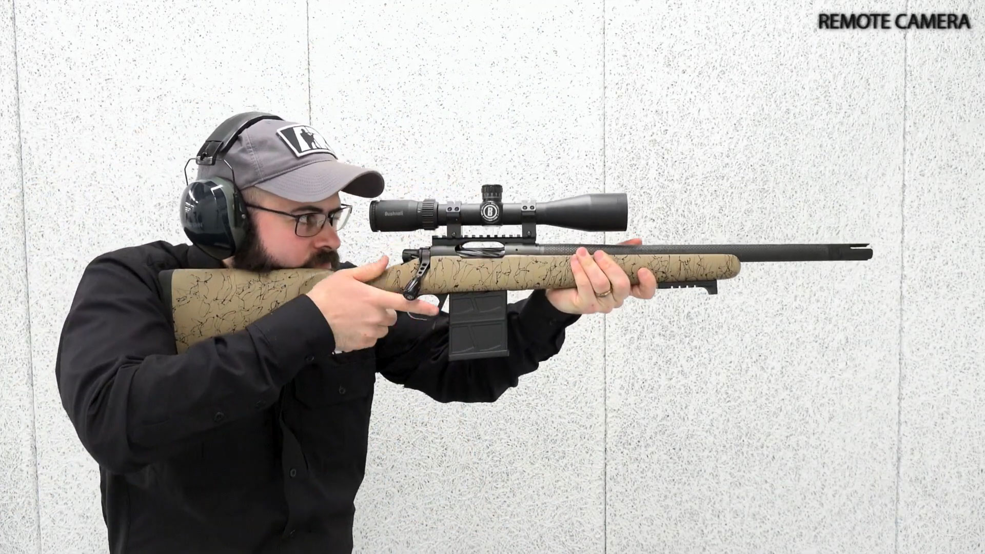 man wearing black shirt ballcap earmuffs glasses shooting rifle bolt-action Christsen Arms rifle scope carbon-fiber barrel white shooting range indoors