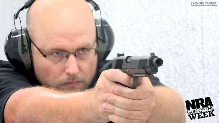 Man wearing protective shooting gear and a black shirt shooting a pistol on a white shooting range.