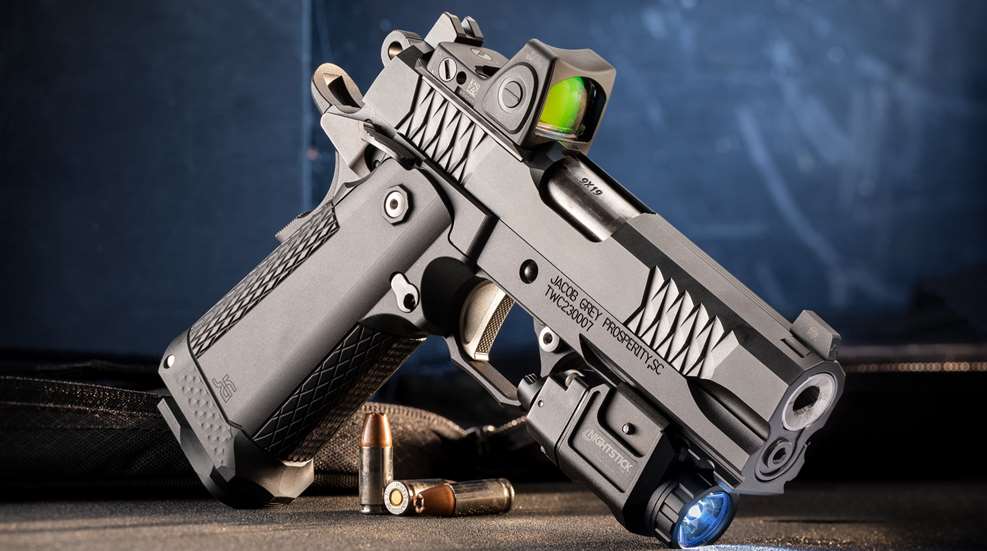 Jacob Grey TWC 9 right-side dynamic angle view pistol with trijicon optic attached nightstick flashlight on blue background shown with ammunition on table