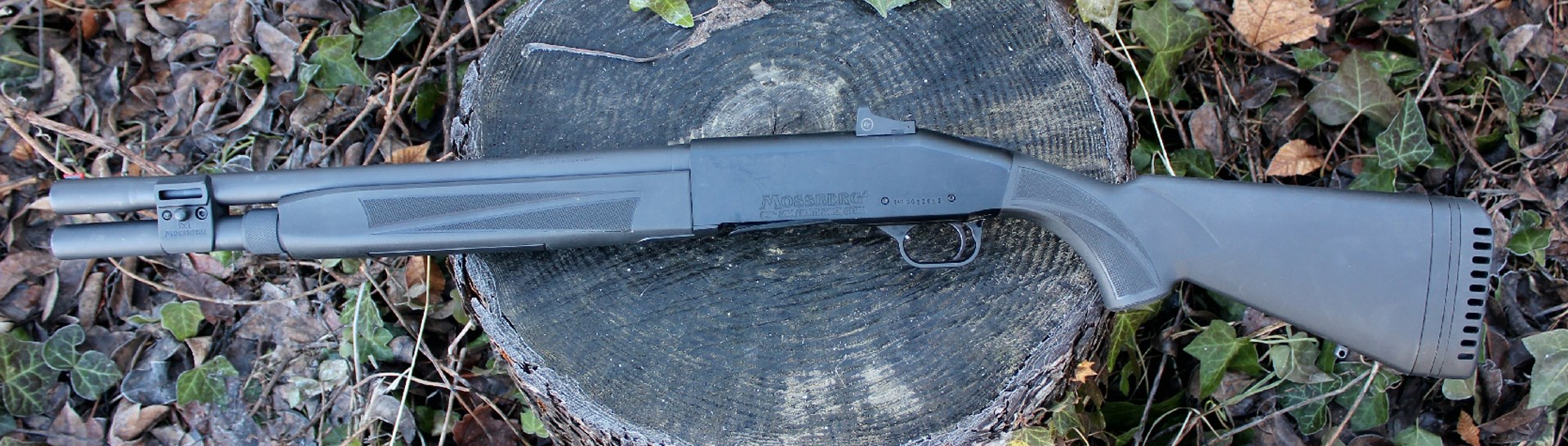 black-colored Mossberg 940 Pro shotgun left-side view resting on log outdoors with leaves sticks and grass