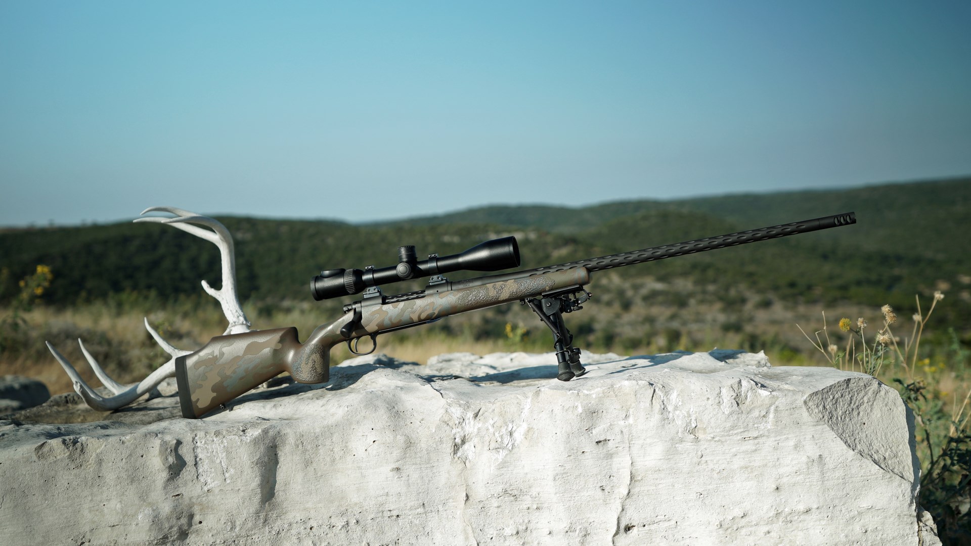 Bolt-action remington m700 custom rifle on rock boulder shown outdoors full sun antlers skull deer background hills ftw ranch