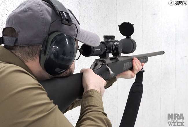 Over-the-shoulder view of rifleman with gray ballcap and green shirt shown in white indoor shooting range holding a black rifle with black Crimson Trace scope and a black sling.