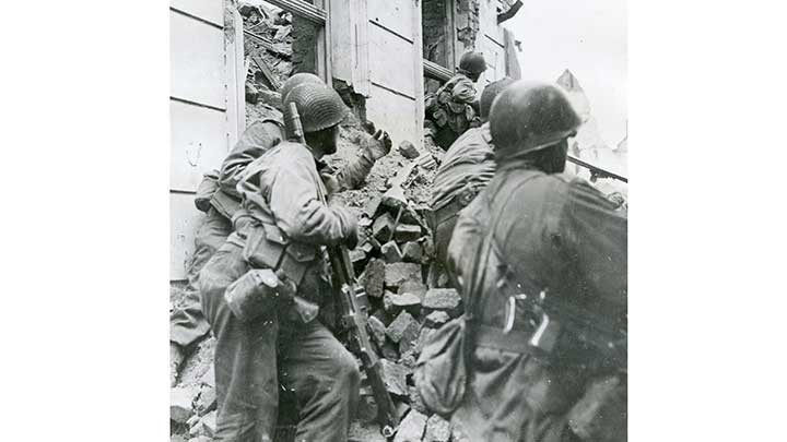 Men of the 104th Infantry Division push past a German roadblock set up in Cologne. The man at center carries a Browning Automatic Rifle. The G.I. to the far right has a M3 “Grease Gun”.