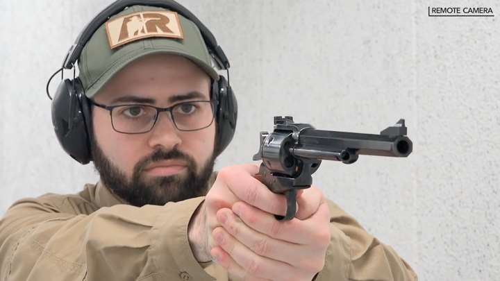 Man wearing a ballcap and protective gear aiming a revolver.