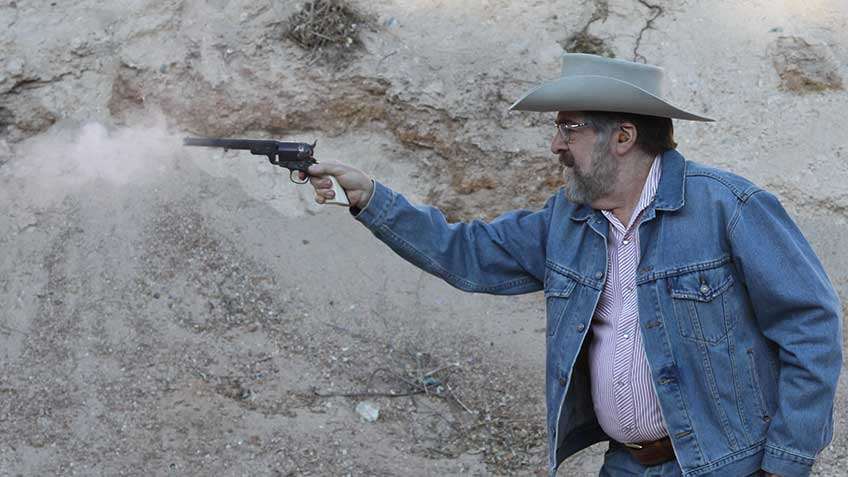 Left-side view of man in cowboy hat and blue coat Shooting the Uberti Wild Bill 1851 Navy Conversion.