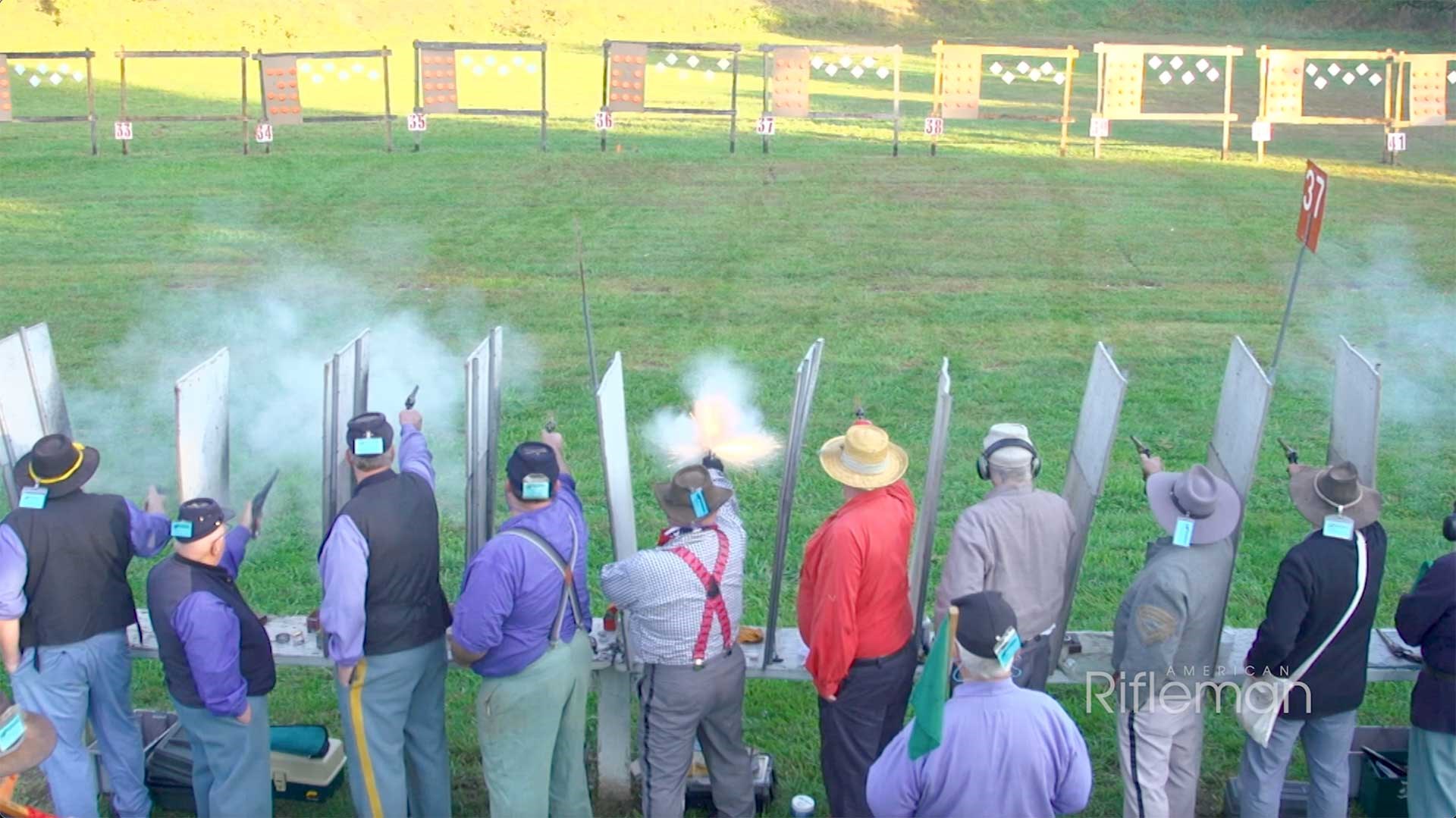 Men on the competition line, aiming revolvers at hanging targets on a green ground.