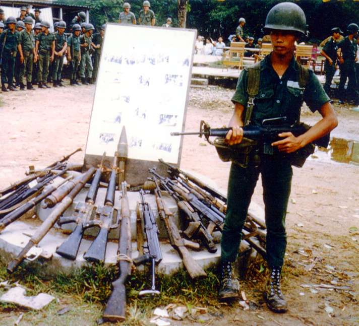 An ARVN trooper guards a typical VC weapons cache—featuring various AK-47s, M16 rifles and a M1 Garand.