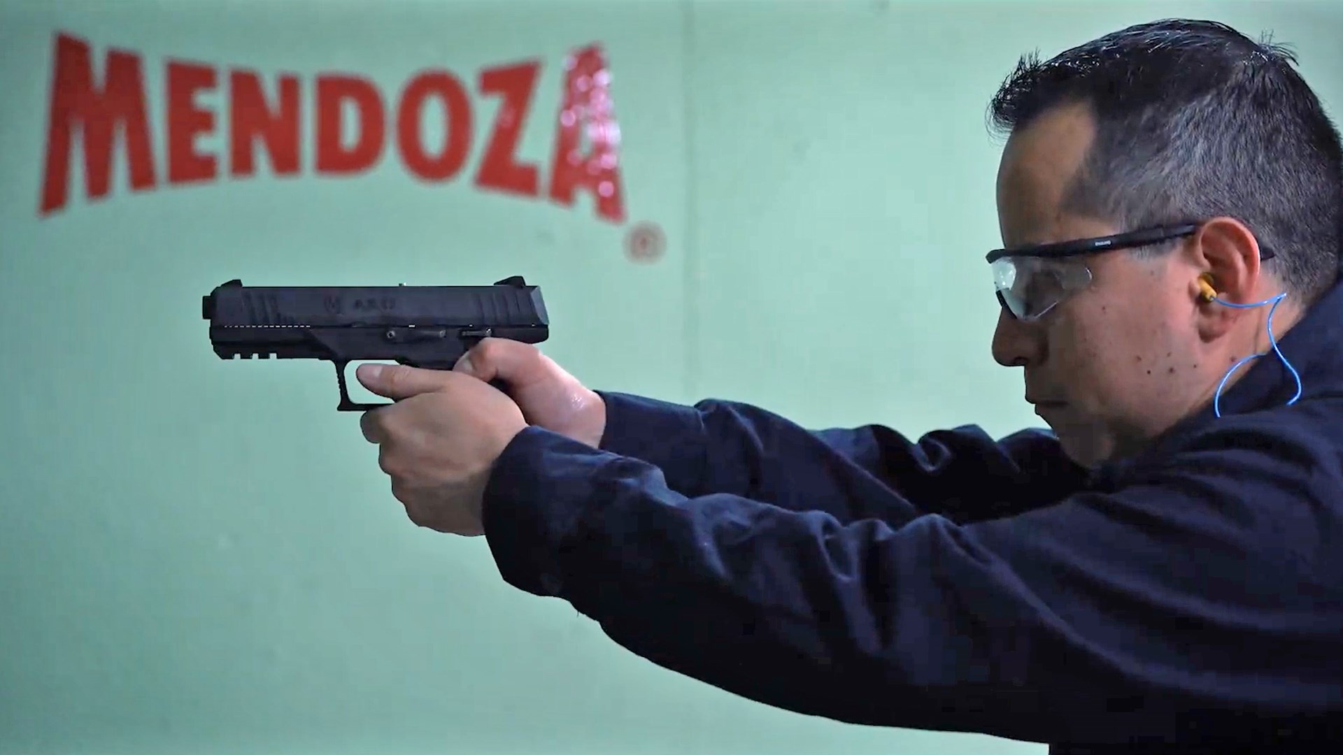 Man aiming a black handgun on an indoor range with the "Mendoza" logo in the background.