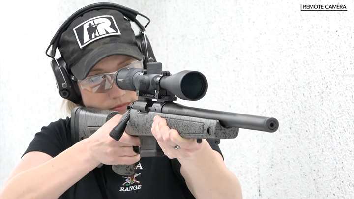 Lady wearing protective shooting gear and camouflage ballcap aiming a rifle on a white shooting range.
