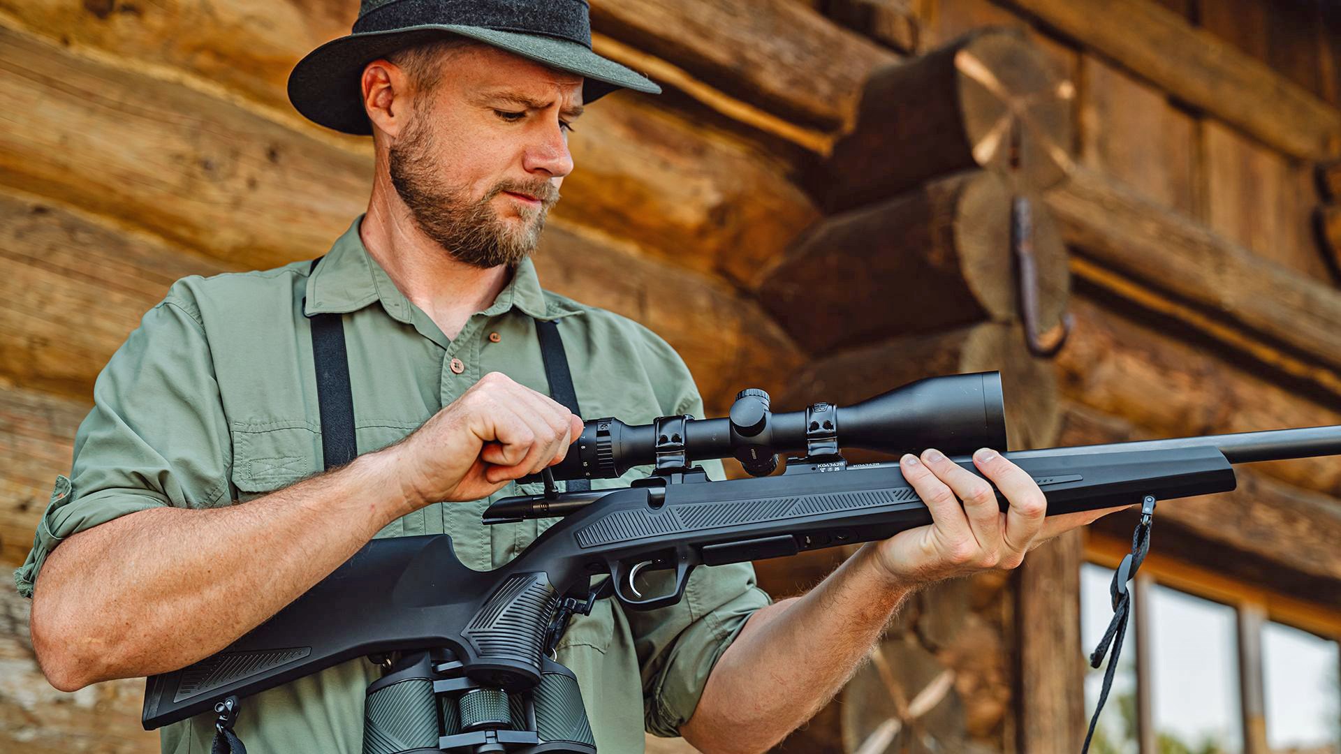 Man in a green hunting shirt operating a CZ 600+ bolt-action rifle in front of a log cabin.