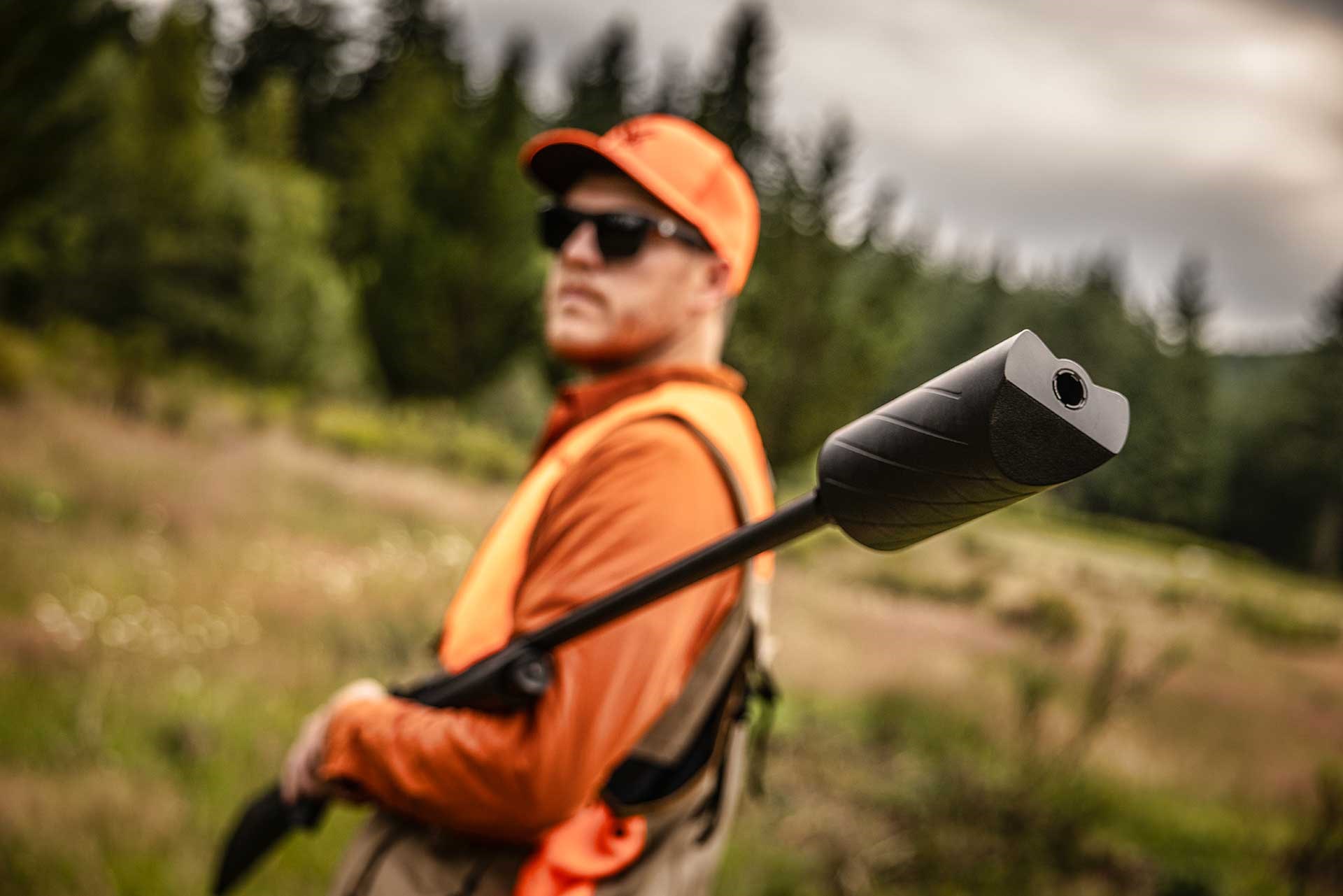 Man in orange hunting gear walking in a field while carrying a shotgun equipped with a Silencer Central Banish 12 suppressor.
