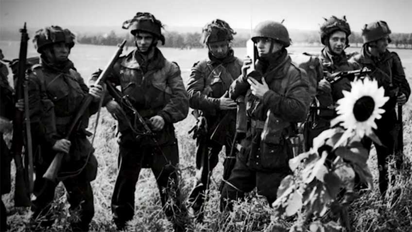 Black and white image of British Paratroopers in Holland, 1944 standing in a field with guns, radio and sunflower growing.