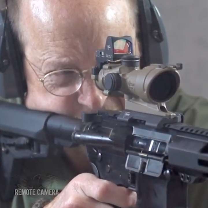 Remote camera image of a man shooting a rifle with a tan scope attached.