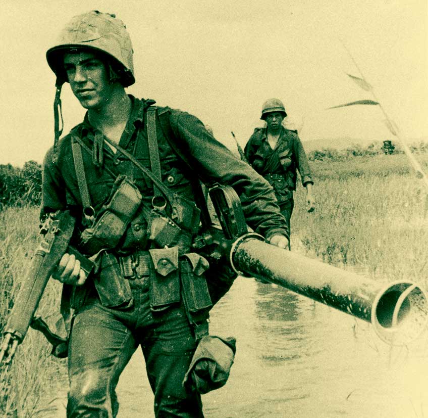An American infantryman wades through swamps in Vietnam, carrying an M14 in his right hand and an M20 bazooka in his left hand.