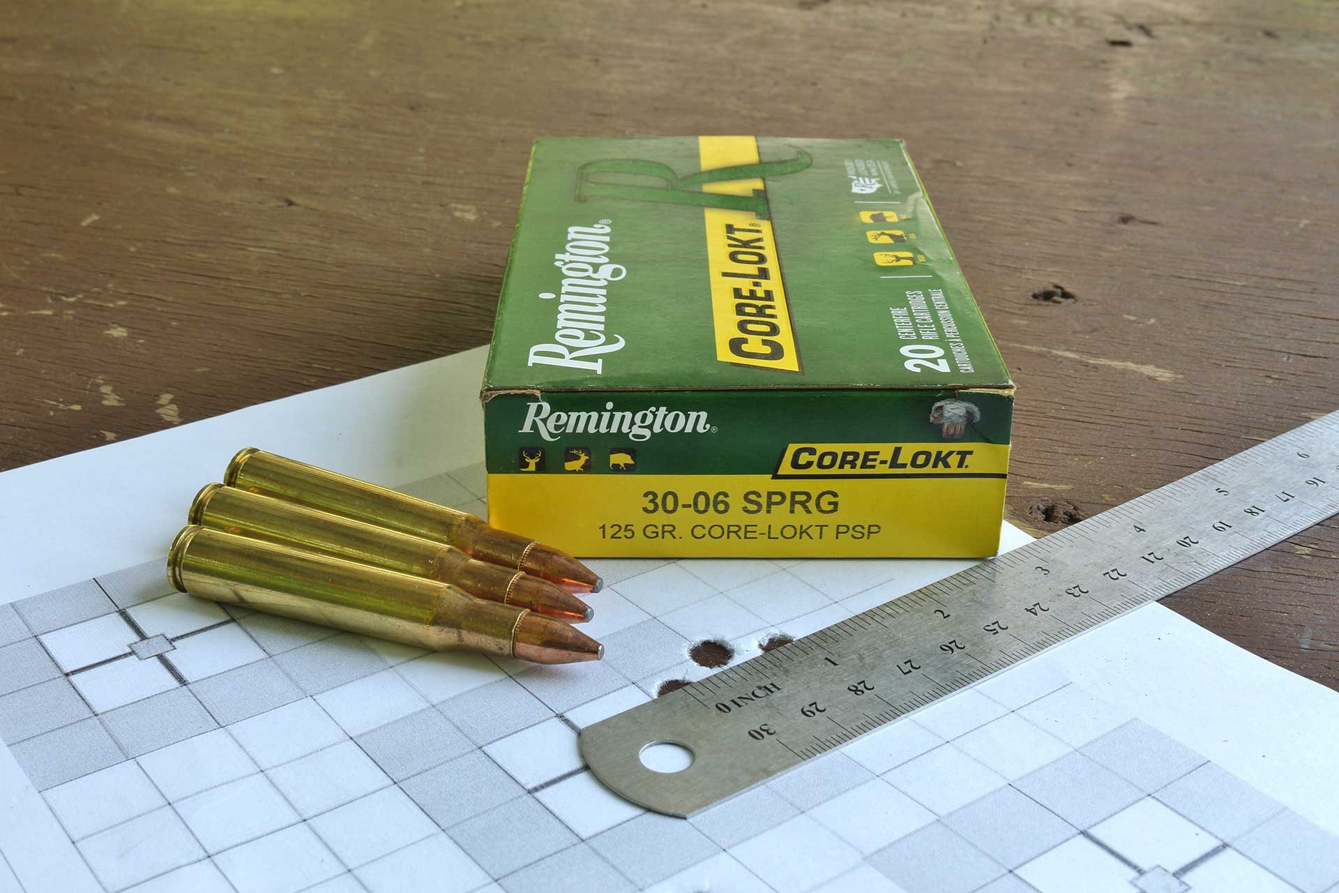 Three .30-'06 Springfield cartridges next to a box of ammunitio and a ruler measuring holes in a paper target.