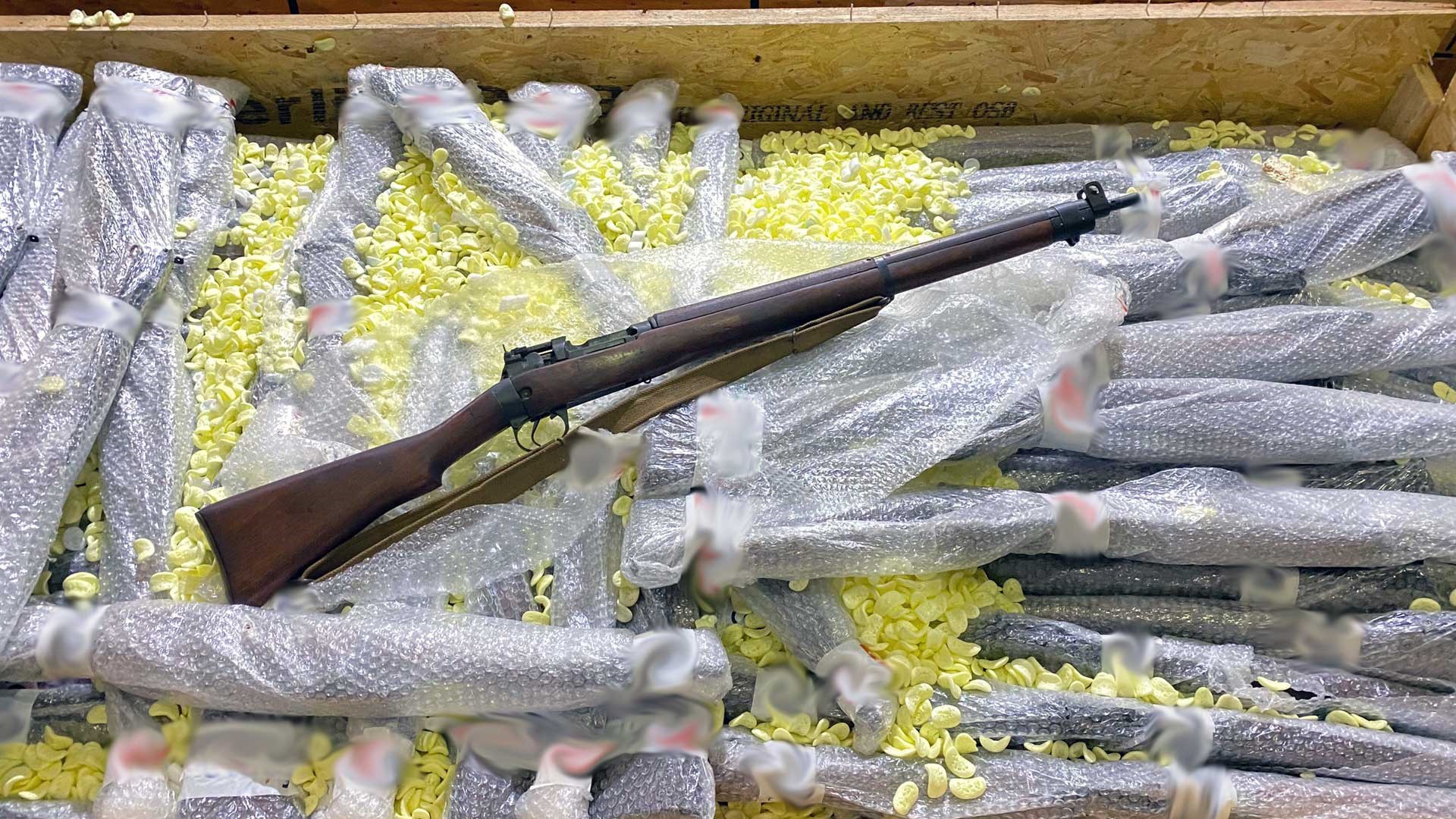 The first rifle is pulled out of its wrap and sits atop the other wrapped No. 4 rifles in one of the shipping containers. Certain identifying information has been obscured at the request of Navy Arms.