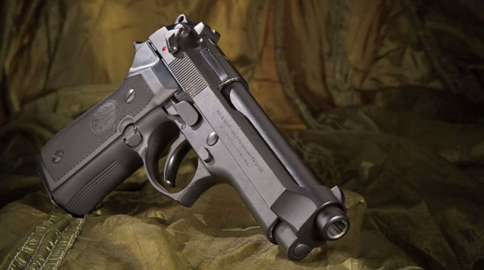 An all-black Beretta M9 propped up against a green, military-like background.