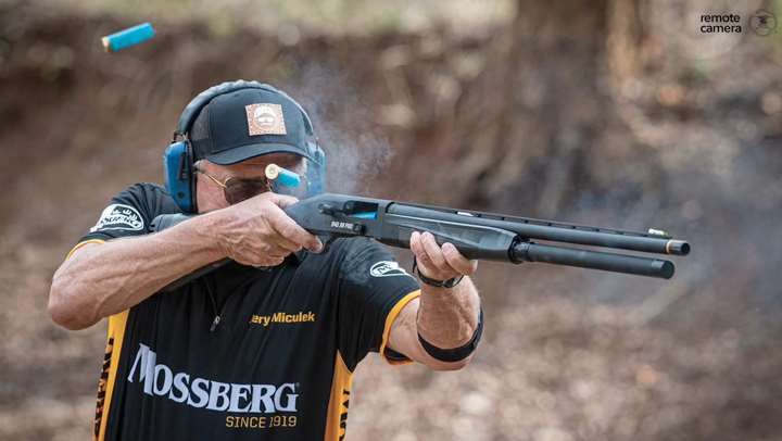 Jerry Miculek shooting a Mosserbg 940 JM Pro shotgun with empty shells flying through the air.