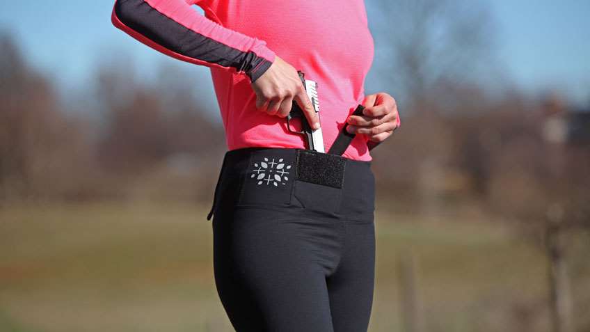 Woman outdoors in pink and red jogging outfit securing a pistol into appendix region.