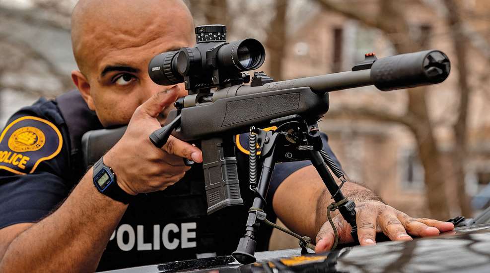 Policeman stretched over vehicle hood sighting bolt-action mossberg rifle with black riflescope