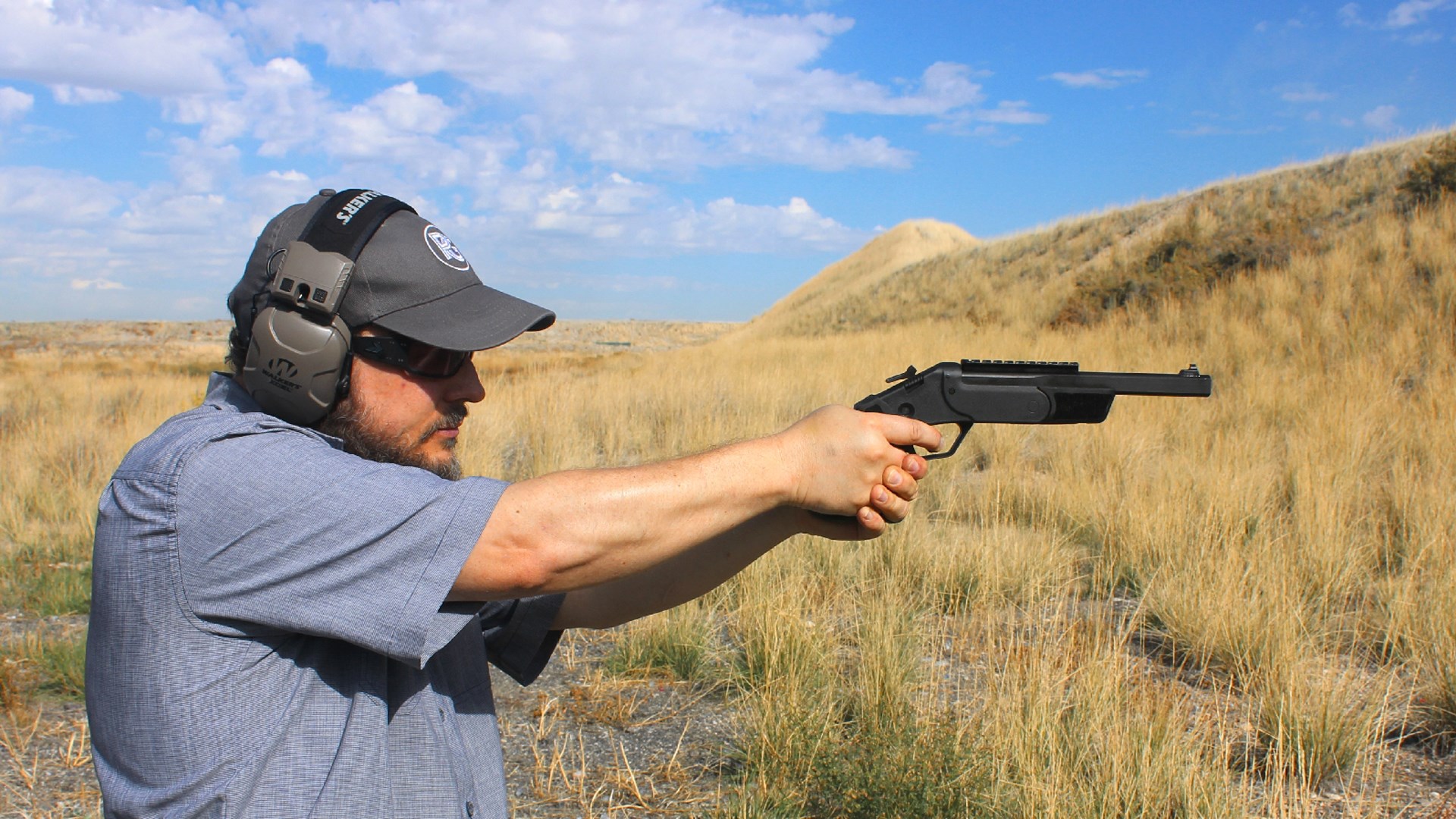Man author wearing hat glasses and earmuffs outdoors holding shooting Rossi Brawler single-shot break-action hinge-action handgun pistol grass mountains blue sky clouds