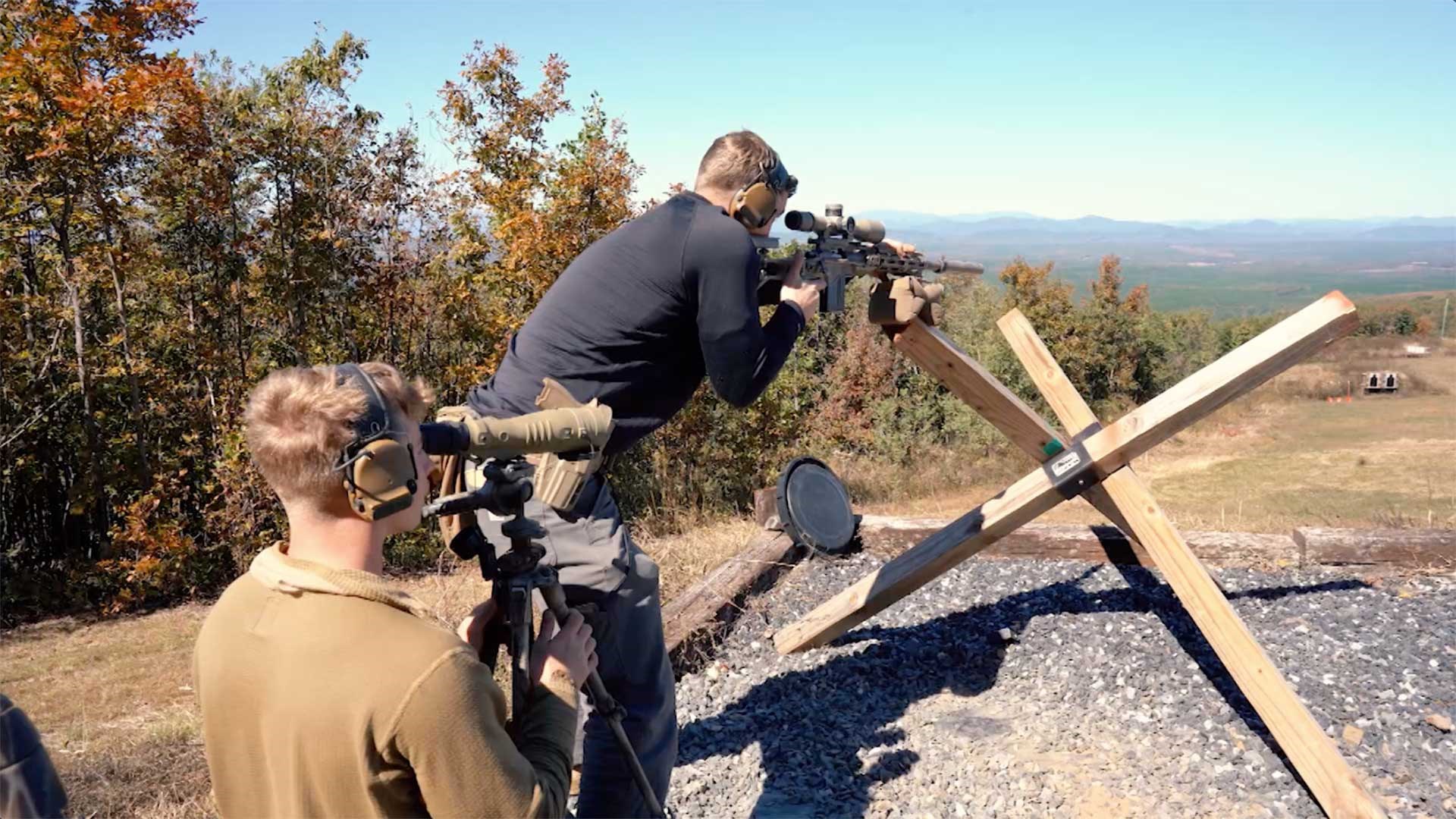 A man wearing a black shirt aims a scoped rifle off a wooden obstacle at the Major Land Sniper Cup shooting match.