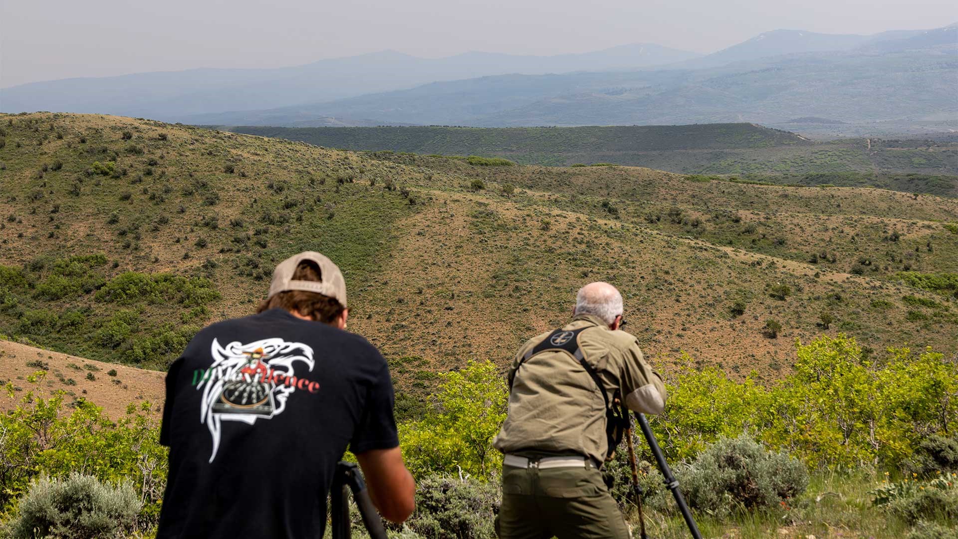 A shooter aims his rifle across mountain ridges while a spotter mounts his spotting scope in the foreground.