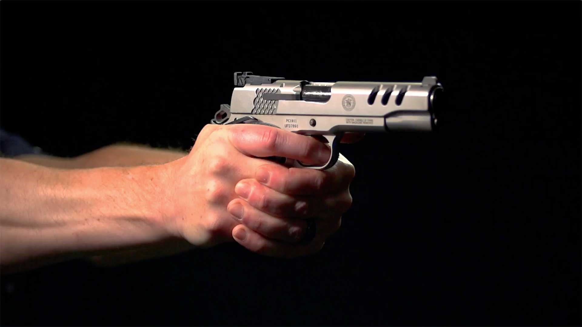 A pair of hands holding a Smith & Wesson Performance Center SW1911 pistol on a dark range.