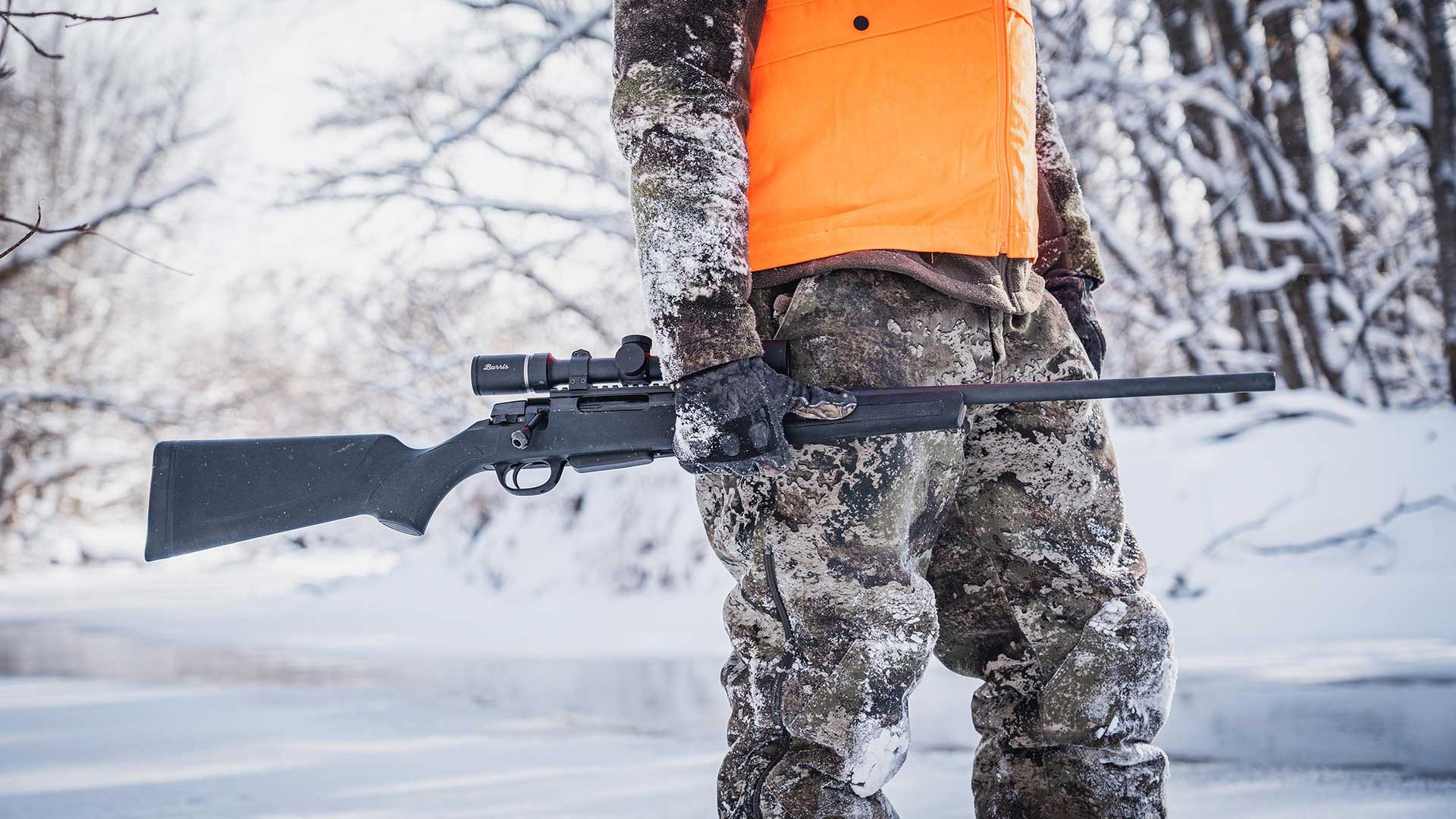 A camouflaged hunter wearing an orange safety vest carries his black Savage 334 bolt-action rifle alongside a frozen river in a snowy winter scene.
