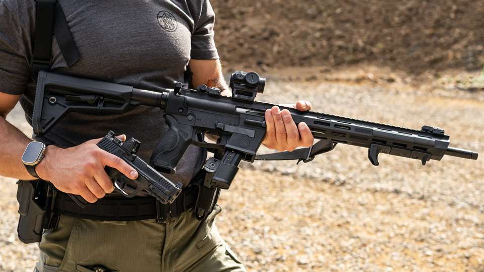 A man holding the Smith & Wesson Response Carbine next to a Smith & Wesson handgun on an outdoor range.
