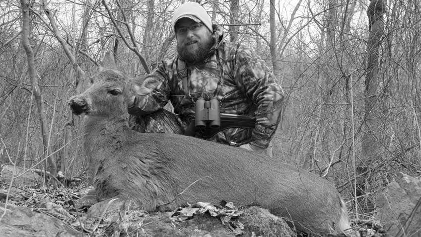 Black and white photo of a beared man dressed in camouflage honoring a whitetail doe harvest with trees, rocks, sticks, mountain.