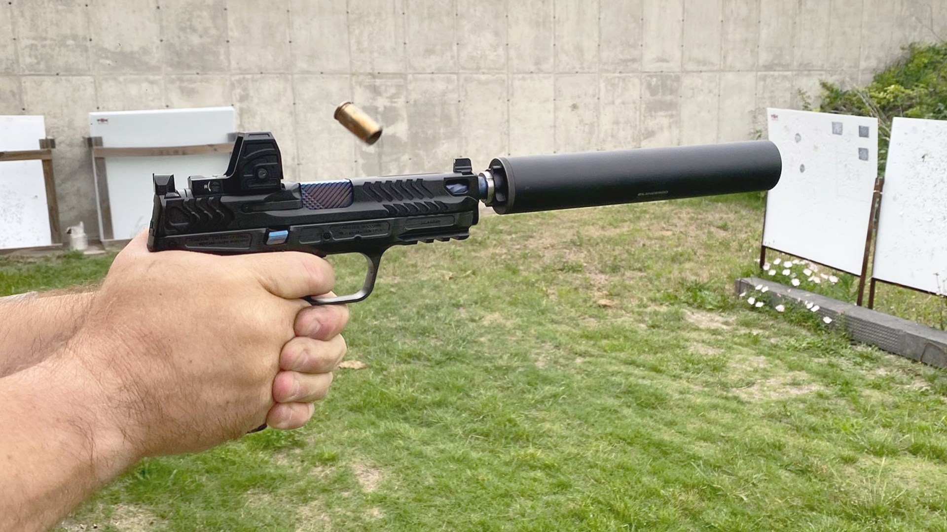 Smith & Wesson Spec Series V M&P 9 Metal HD held in hand firing 9 mm Luger brass flying from gun shown with black suppressor and red-dot optic