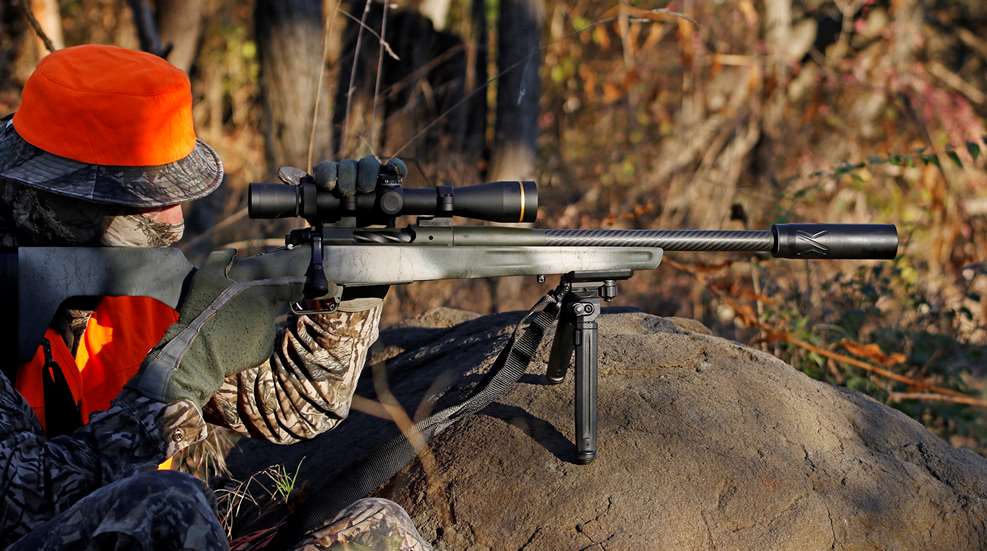 Camouflaged hunter with blaze orange safety colors perched on a rock with bolt-action springfield armory redline rifle carbon-fiber barrel suppressor outdoors