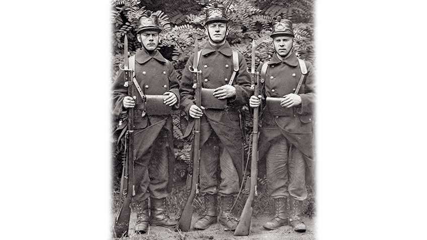 Black and white image of three soldiers in uniform, helmuts on and rifles standing in front.