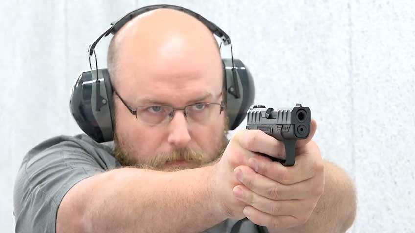man in gray shirt with earmuffs and glasses shooting a black pistol
